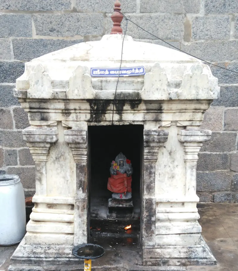 A small white stone shrine with a Tamil name board, holding a dark image in red cloth, with a lamp burning below.