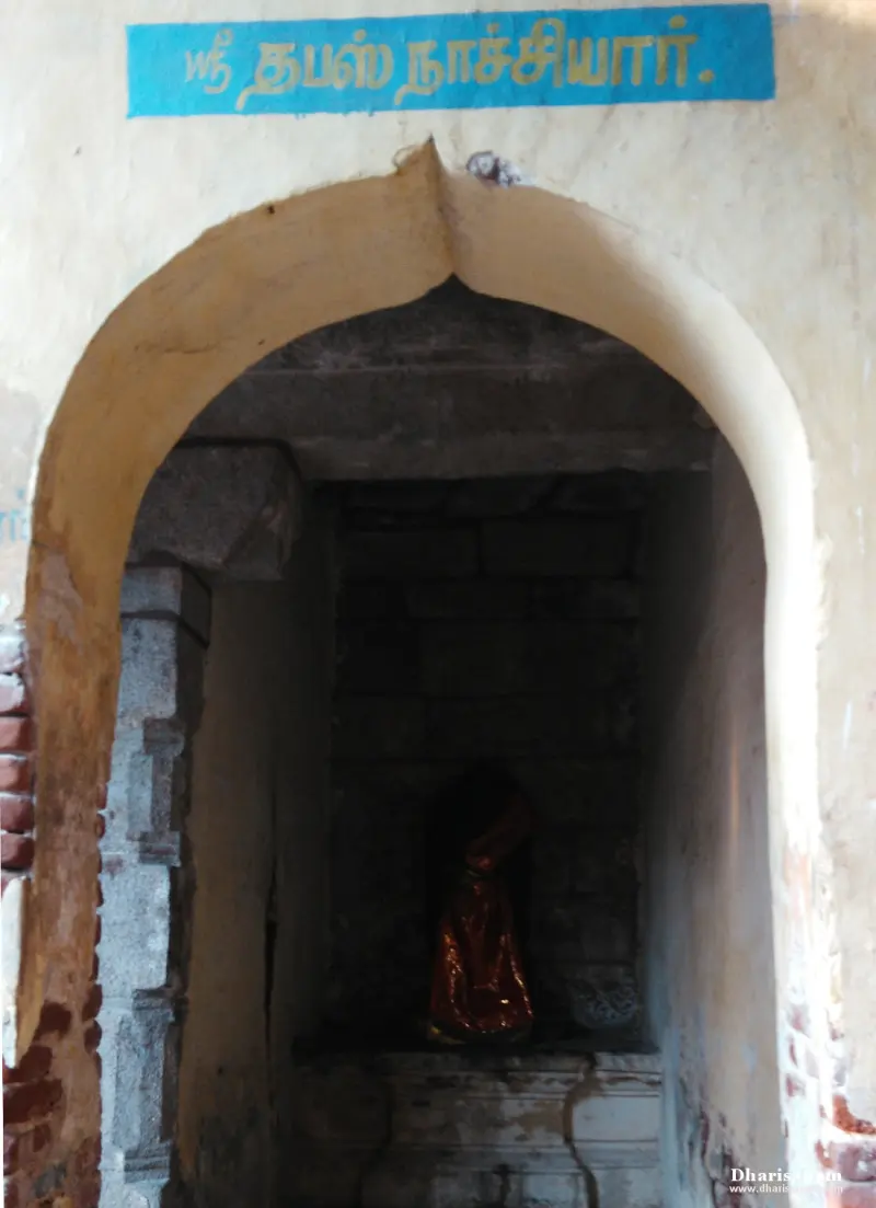A small arched shrine with a figure in a red cloth inside, under a blue Tamil name board