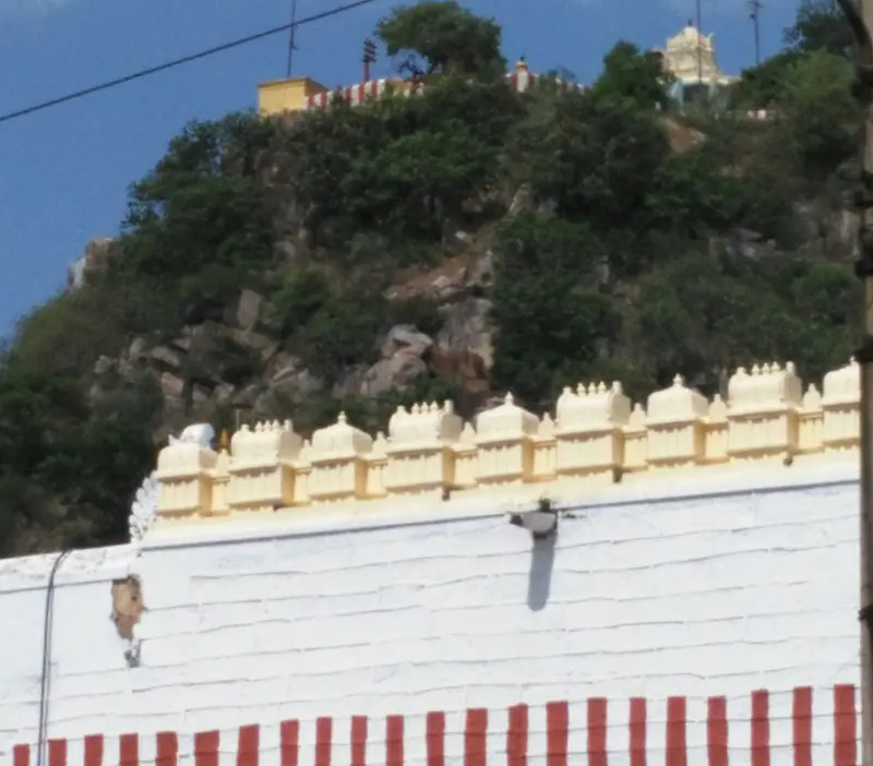 A white wall with red stripes and a row of cream parapets, a rocky wooded hill behind with small shrines at the top.