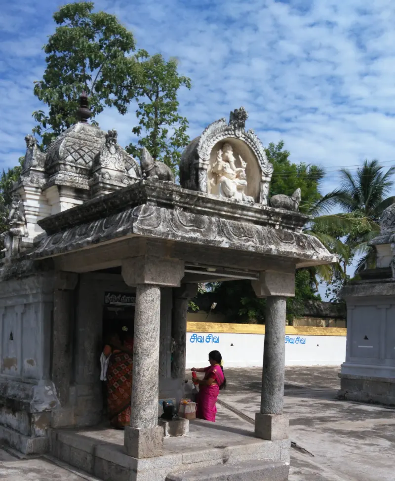 A stone shrine porch with two pillars and an elephant-headed deity carved above, with a woman in pink offering worship.