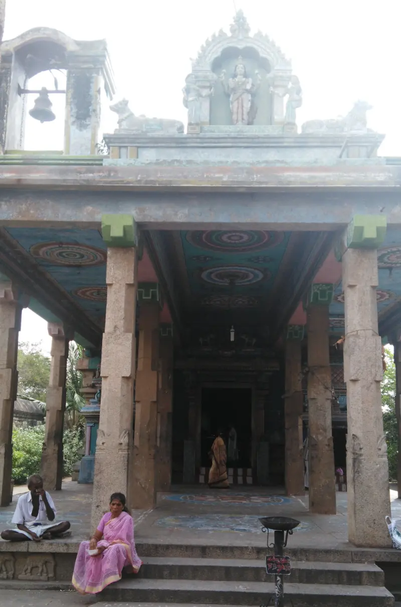 An open mandapam with stone pillars and a painted ceiling, a bell at the left and a goddess figure above