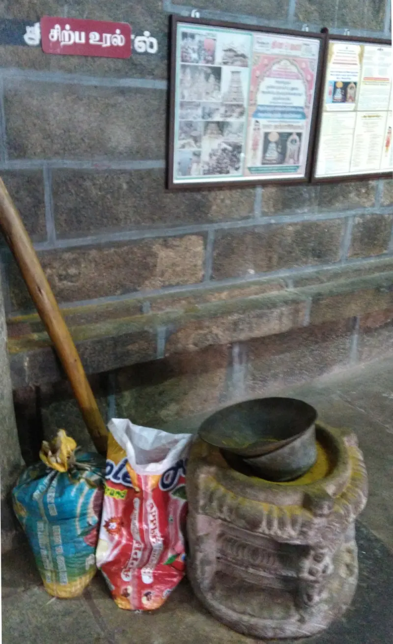 A carved stone grinder with a metal bowl beside a wall board reading Sirpa Ural, with sacks next to it