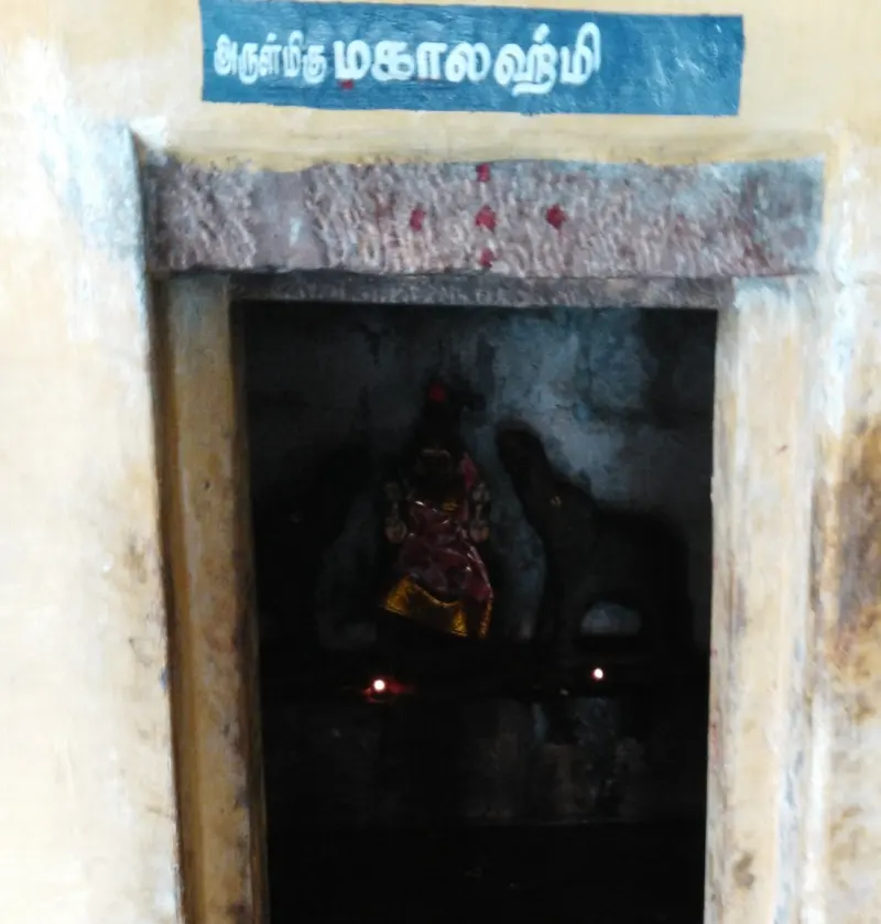 A dark shrine opening with a blue Tamil name board; a small idol in a red and gold sari is faintly seen inside