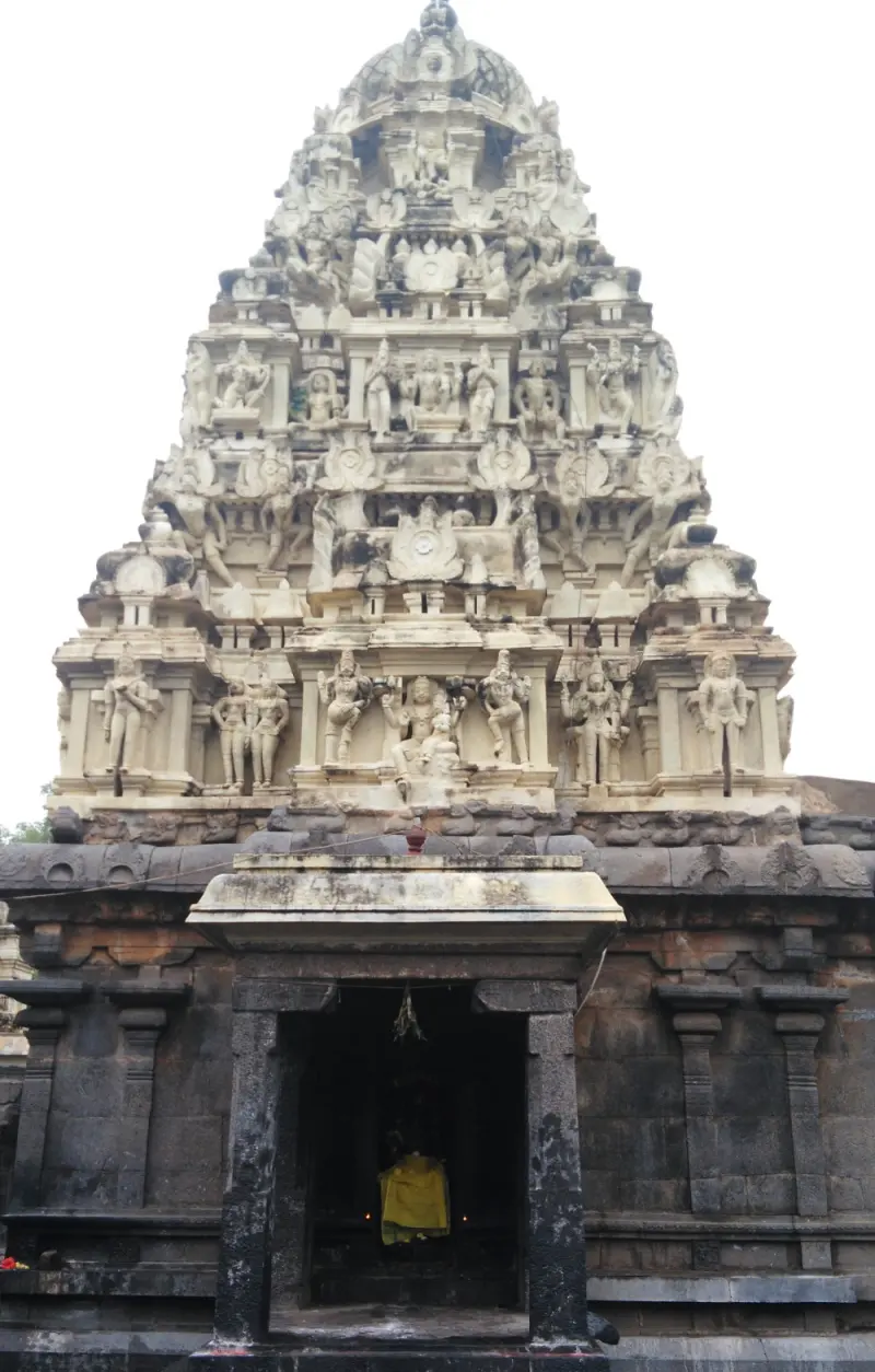 A tall tower covered with pale carved figures above a dark stone wall and a doorway with an image in yellow cloth.