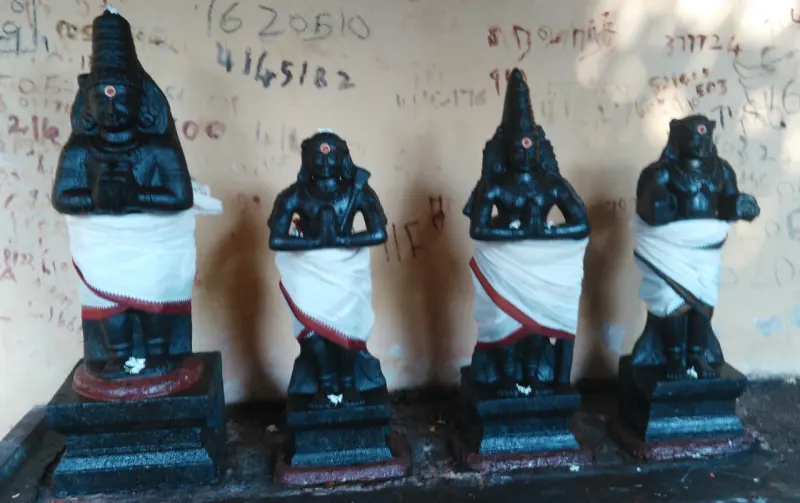 Four small black stone idols in white cloth stand in a row on stone pedestals against a wall.
