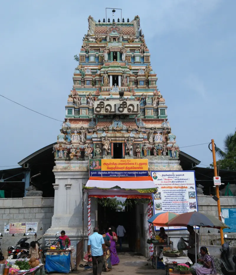A tall painted gopuram above the entrance, with devotees and flower sellers on the street below.