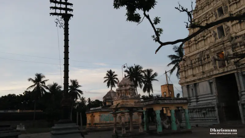 A tall flagstaff beside a small pillared shrine, with a tower at the right edge and palms behind