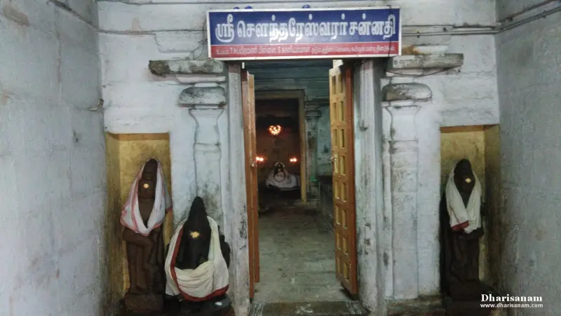 Open wooden doors under a Tamil name board, with a dark idol dressed in cloth standing on each side