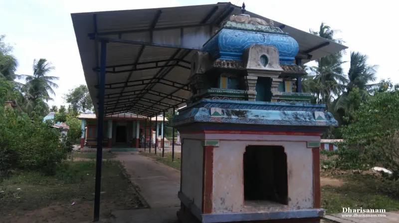 A small blue-topped shrine with an open niche beside a long tin-roofed walkway leading to a hall