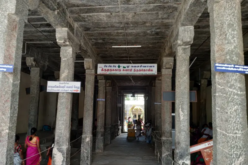 A long stone hall with rows of pillars and a name board hanging overhead, with devotees at the far end and sides.
