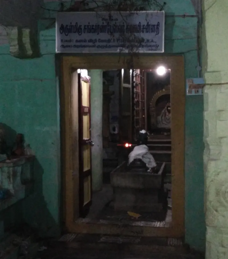 A lit doorway under a name board, with a lingam seen inside and a cloth-covered pedestal in front.