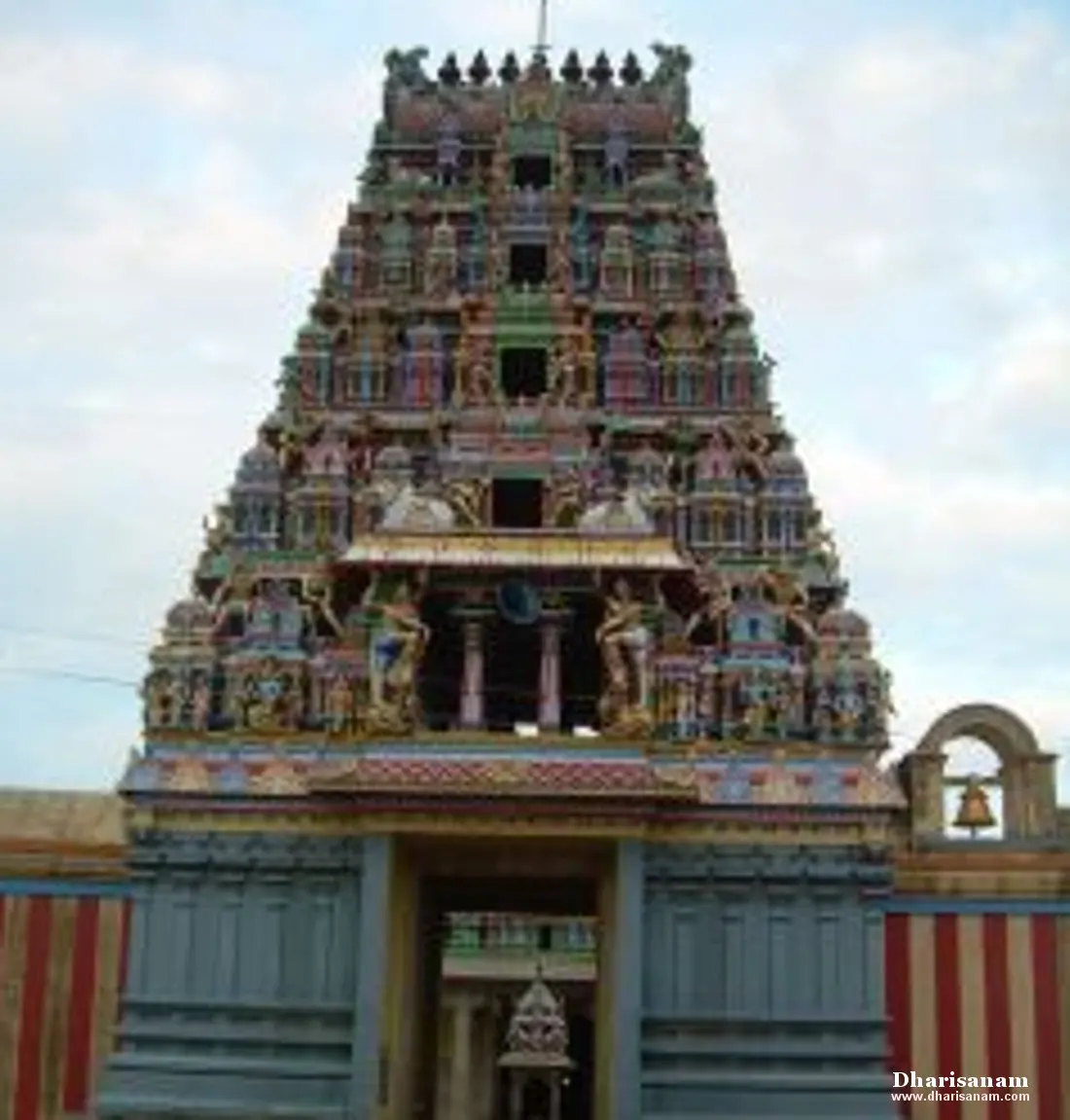 A tall many-tiered gopuram covered in painted figures, above a dark entrance; a bell arch to the right.