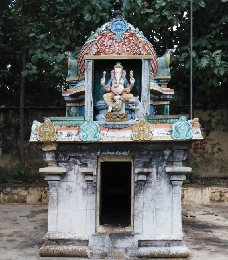 A small stone shrine with a painted Vinayakar figure and mouse beneath a domed roof; Tamil lettering above the door.