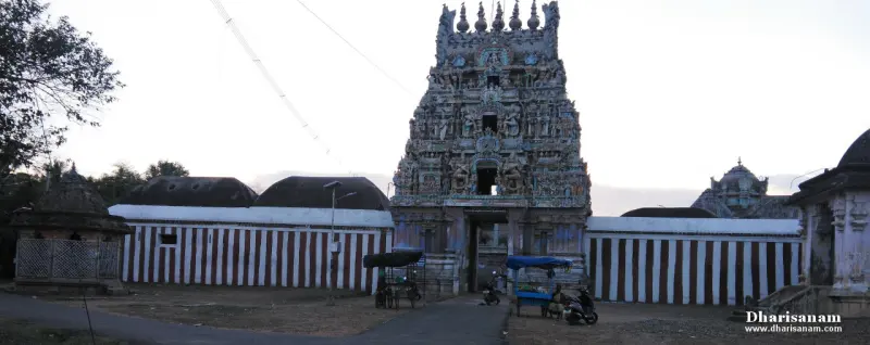 A carved gopuram rises above a gateway with red and white striped walls on both sides, seen at dusk from the road