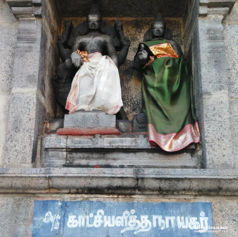 Two stone idols, one in a white cloth and one in green and gold, in a niche above a blue Tamil name board.