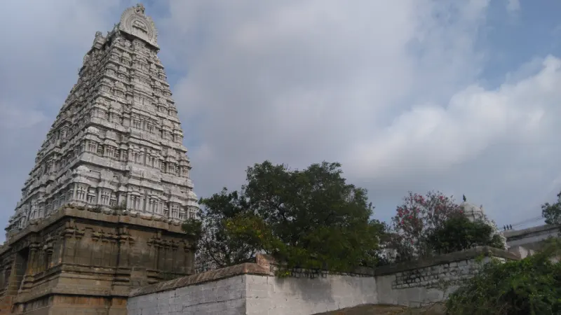 A tiered gopuram at the left, a whitewashed stone wall and green trees under a cloudy sky