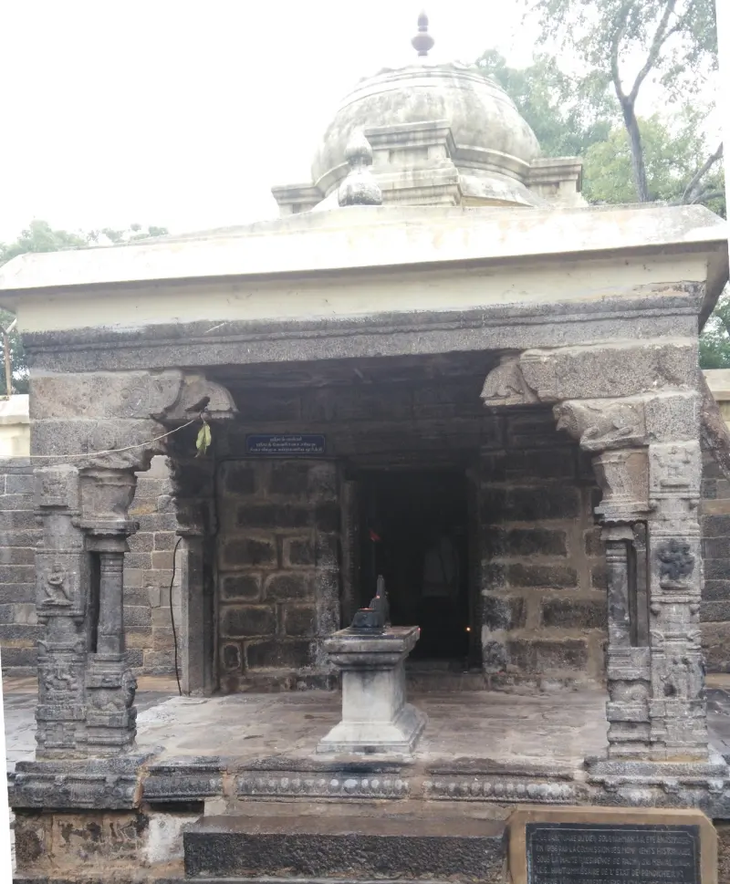 A small open stone shrine with carved pillars and a round domed roof, and a stone pedestal before the dark doorway.
