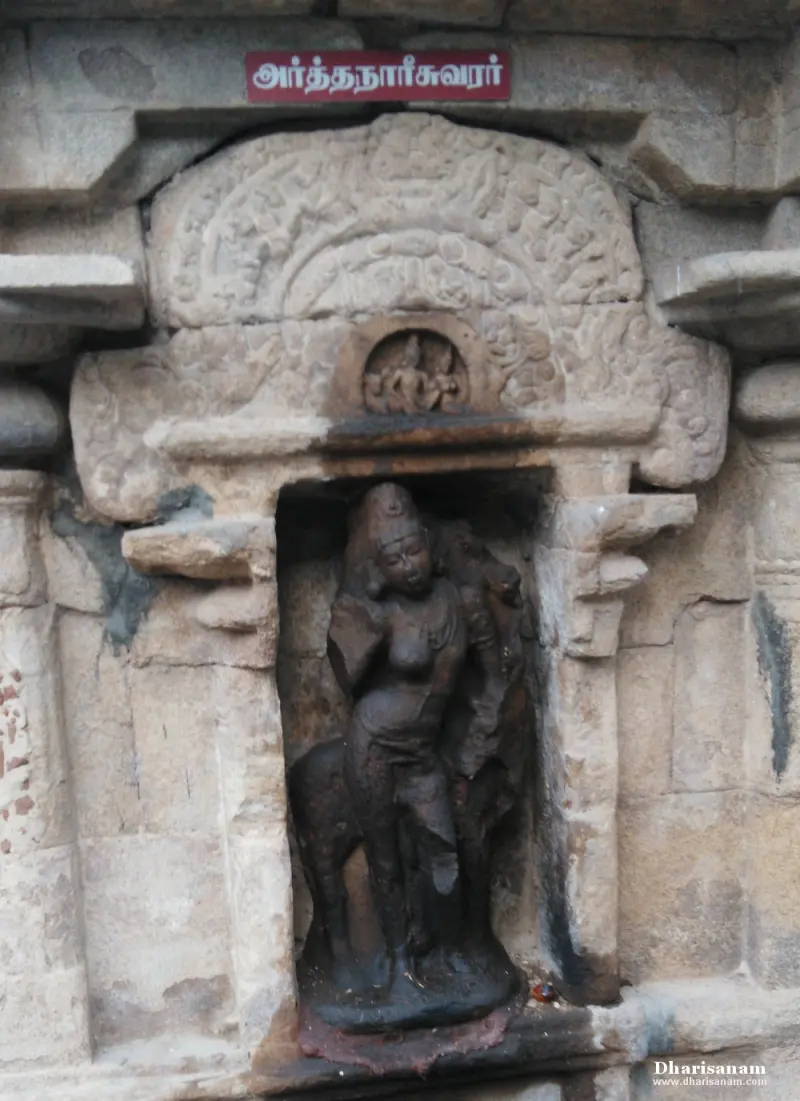 A weathered stone figure of Ardhanareeswarar in a carved niche, with a red Tamil name board above.