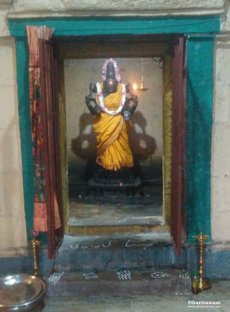Black stone goddess idol in a yellow sari standing in a niche with teal frame and lamps at the foot