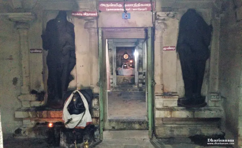 A Vinayakar idol at the left of the sanctum doorway, a tall guardian figure on each side, the sanctum lit within