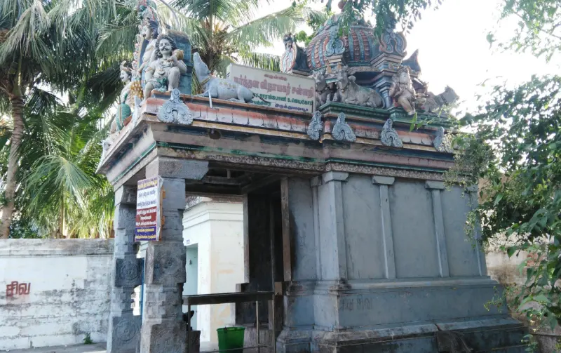 A grey stone gateway with a signboard and stucco figures on top, with palms and trees around it