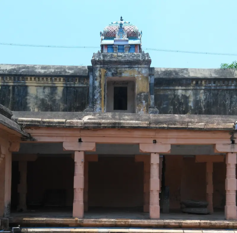 A pillared corridor in front of a weathered wall, with a small painted tower above its roof.