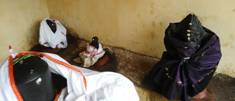 A lingam and two small stone figures draped in white and purple cloth, against a yellow wall