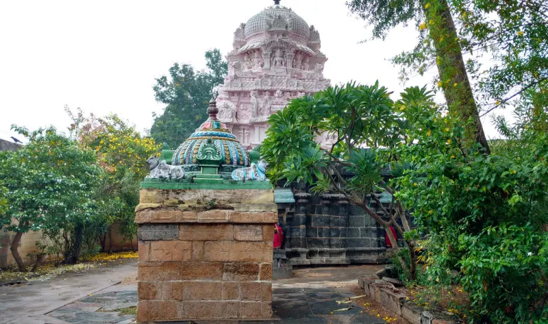 A small stone shrine with a green-roofed top, and a pink tower rising behind trees.