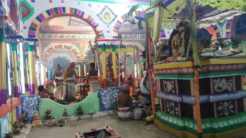 Priests and devotees seated and standing in a brightly painted hall with flower garlands and a decorated platform.