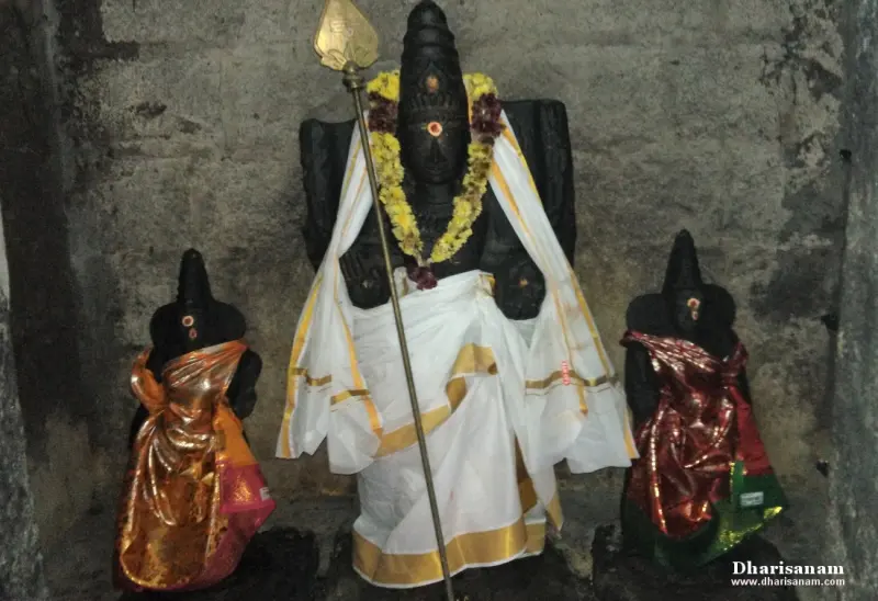 A dark stone Murugan with a spear, flanked by two smaller consort idols draped in bright silk, with garlands.