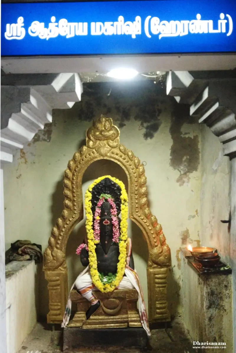 A dark seated idol with flower garlands under a golden arch, beneath a blue name board