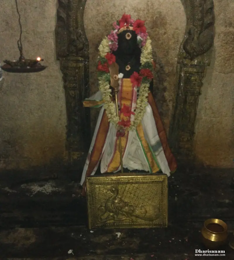 A standing idol with a staff, in garlands and cloth, above a brass plate showing a peacock.