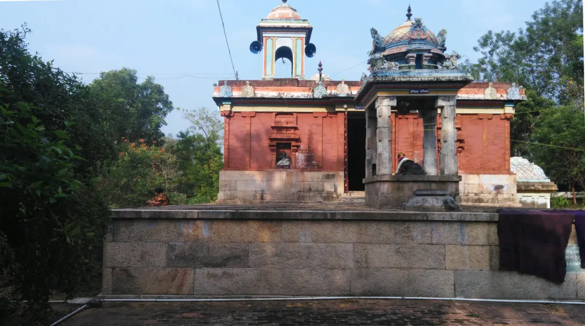 A red-walled temple with a small mandapam in front, standing on a raised stone platform among trees.