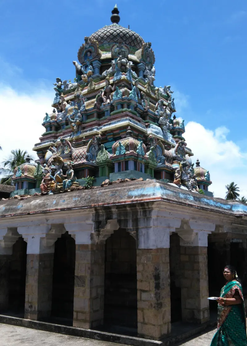 A tiered, sculpted tower above a stone pillared hall, with palm trees behind and a woman at right.