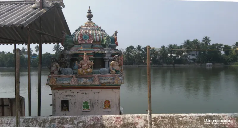 A small painted shrine with a seated figure at the edge of a wide pond, palm trees on the far bank