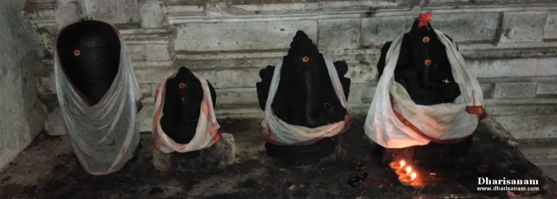 Four dark stone idols draped in white cloth, with small lit lamps in front of them