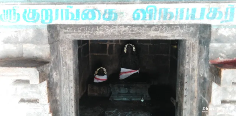 A small stone shrine with two garlanded idols inside, under a blue Tamil name board