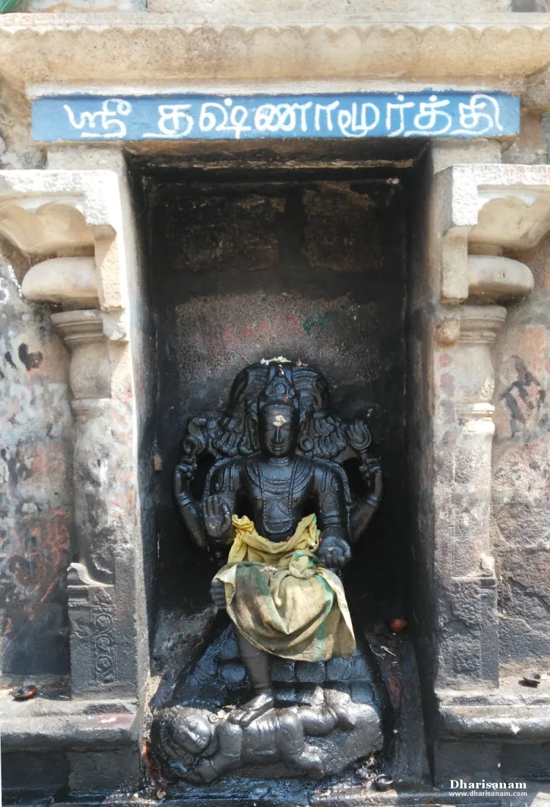 A black stone Dakshinamurthy in a wall niche beneath a blue board with his name in Tamil