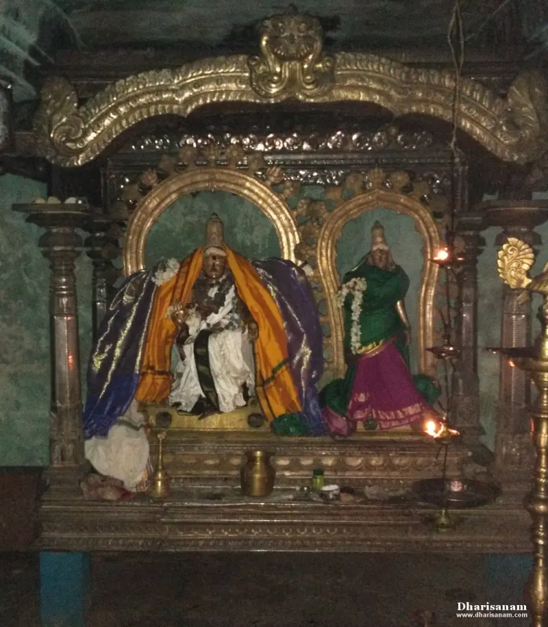 A seated, silk-draped god and a standing goddess in garlands inside a gilded frame with lit lamps