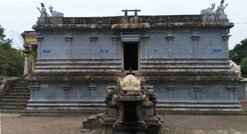 A grey stone temple building with steps at the left, a small shrine in front and statues on the roof corners.