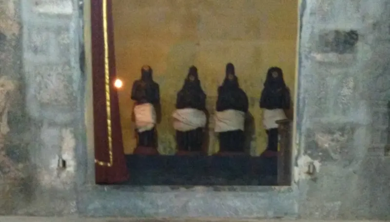 Four small dark idols stand in a row in a lit niche in a stone wall, with a lamp at the left.