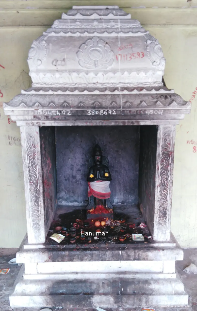 A small black stone figure with folded hands inside a white stone shrine, with oil lamps before it