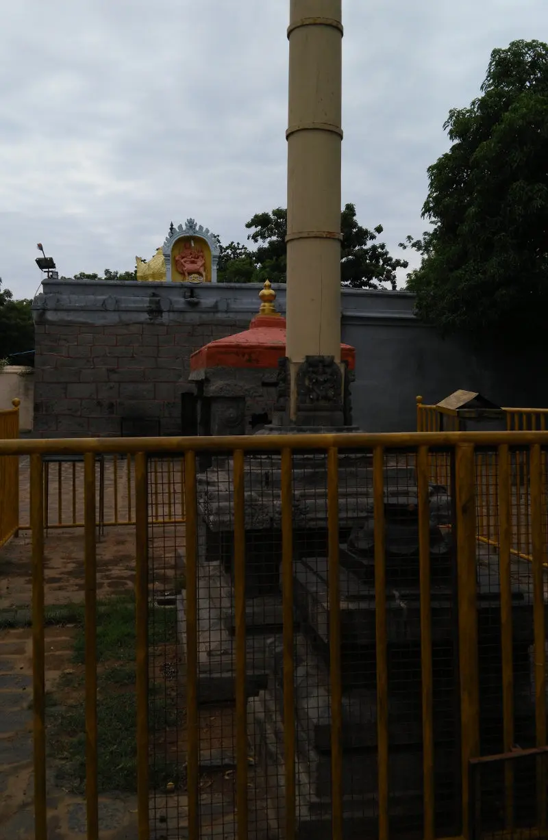 A tall flag post rises behind a yellow railing, with a small red-roofed shrine and a stone wall behind.