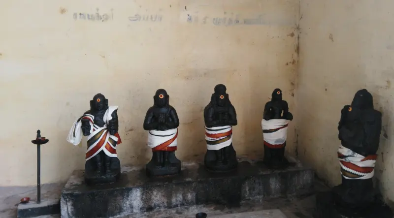 A row of black stone figures wrapped in cloth, standing on a low platform against a plain wall