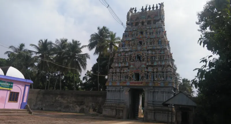 A gateway tower with many painted figures, with palms and a small pink building beside it