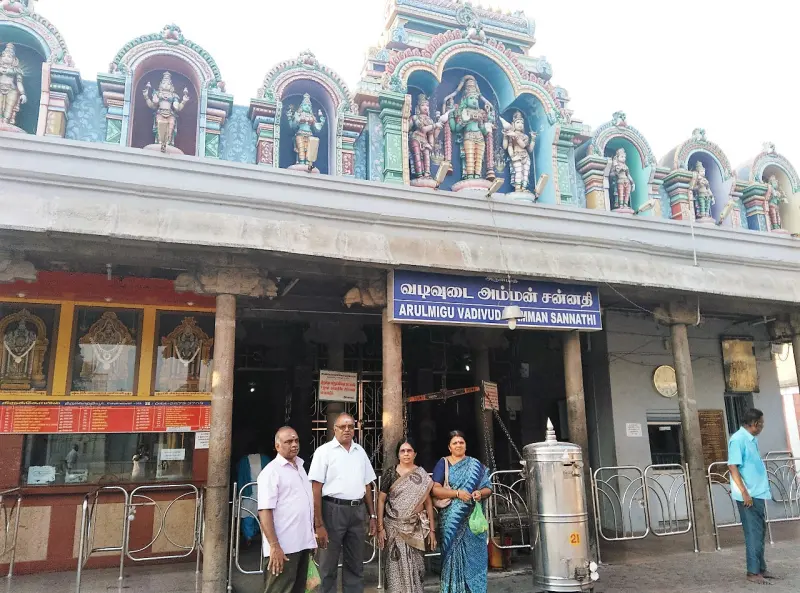 A shrine front with a blue name board and painted figures on the roof, with visitors standing outside