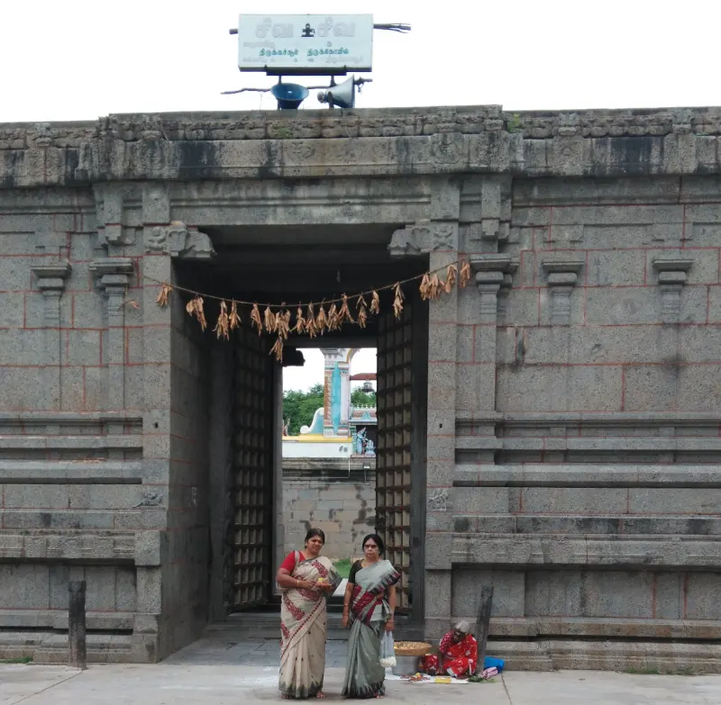 A plain stone gateway with a garland of dried leaves across it and two women standing in front.