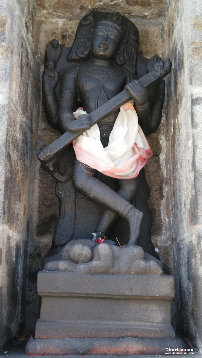 A dark stone goddess holding a long weapon, with one foot raised, standing above a reclining figure.