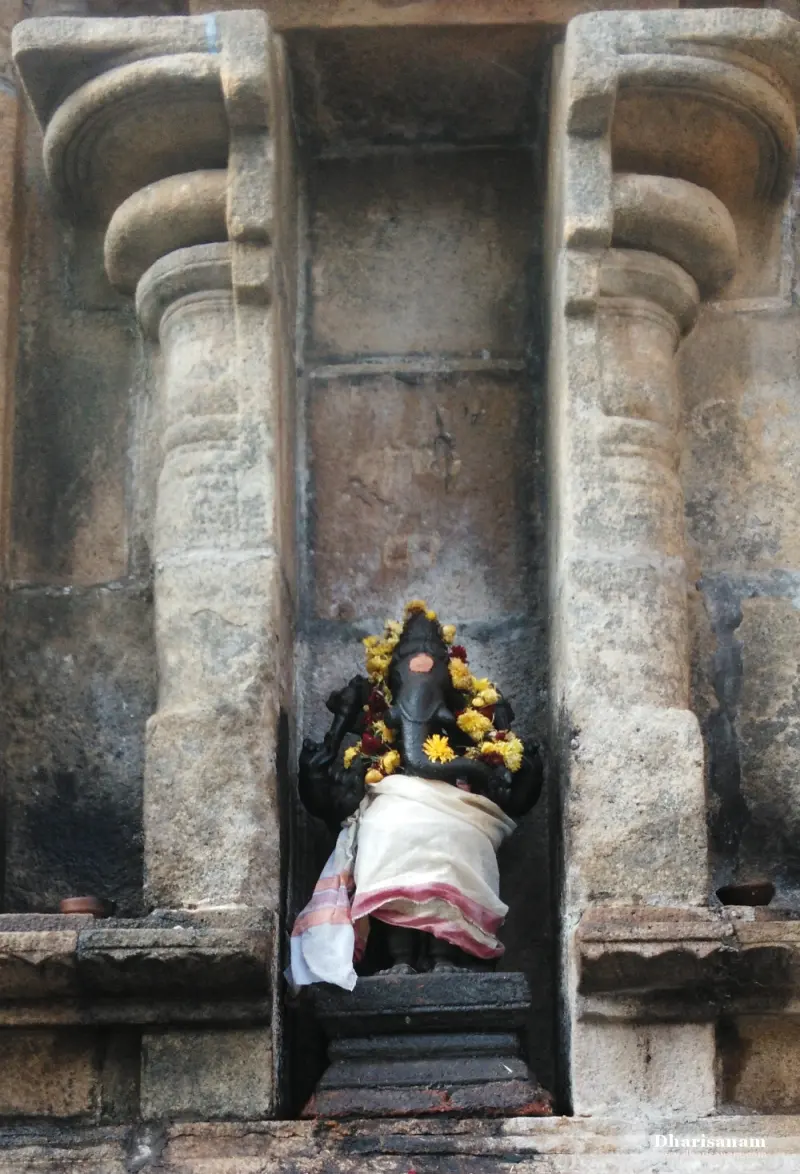 A dark Vinayakar idol with a flower garland and white cloth stands in a stone niche between two pillars