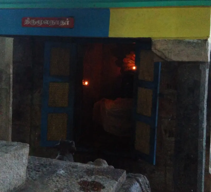 A small dark shrine with open blue doors and a red Tamil name plate; an oil lamp glows inside.
